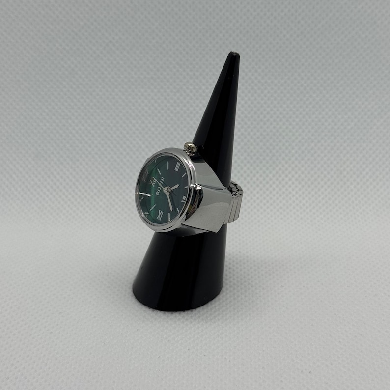 May include: A silver ring watch with a dark green face and silver hands. The watch has a round face with Roman numerals and is displayed on a black cone-shaped stand. The band is expandable.