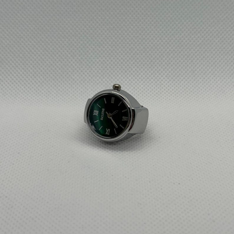 May include: A silver ring watch with a dark green face and black Roman numeral hour markers. The watch face has the brand name "BOZHI" printed on it. The ring is a unique accessory.