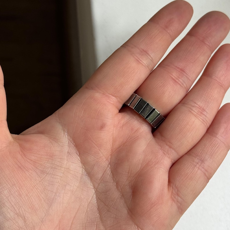 May include: A silver-toned ring with a rectangular block design. The ring is worn on a finger, showcasing its geometric pattern and metallic sheen. The ring's design is modern and minimalist.