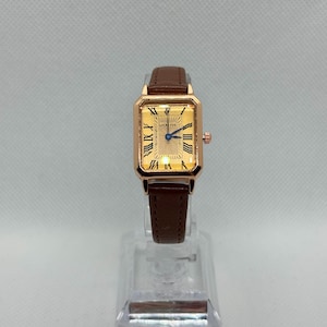 Può includere: Un orologio da polso rettangolare color oro con cinturino in pelle marrone. Il quadrante dell'orologio presenta numeri romani e lancette blu. L'orologio è etichettato "LUCKY FOX".