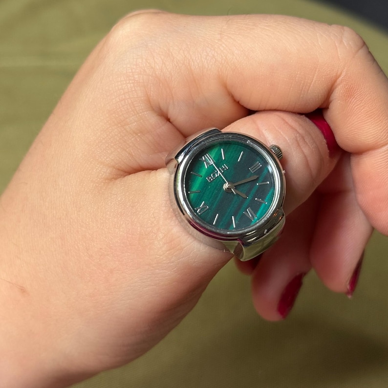 May include: A silver ring watch with a green face and Roman numeral hour markers. The watch face has a marbled appearance. The ring is worn on a finger.