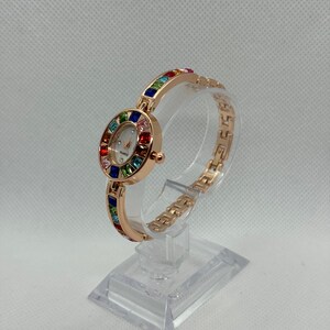 May include: A rose gold quartz watch with a white face and a bezel and band adorned with colorful gemstones. The watch is displayed on a clear stand, showcasing its elegant design. The watch band is approximately 7 inches long.