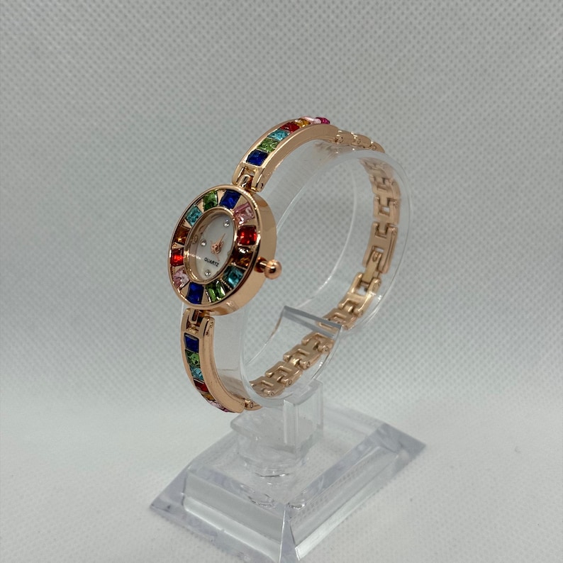 May include: A rose gold quartz watch with a white face and a bezel and band adorned with colorful gemstones. The watch is displayed on a clear stand, showcasing its elegant design. The watch band is approximately 7 inches long.