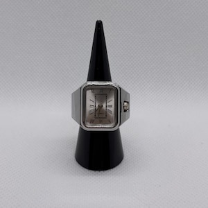 May include: A silver-toned ring watch with a rectangular face. The watch face has Roman numeral hour markers and a quartz movement. The ring is displayed on a black cone-shaped stand against a white background.