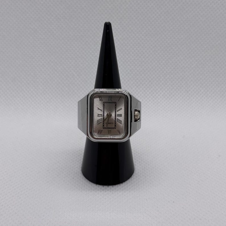 May include: A silver-toned ring watch with a rectangular face. The watch face has Roman numeral hour markers and a quartz movement. The ring is displayed on a black cone-shaped stand against a white background.