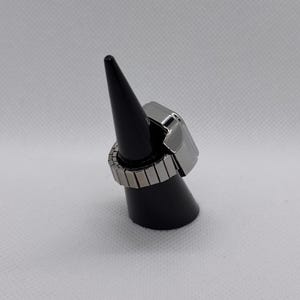 K&ouml;nnte beinhalten: Ein silberfarbener Ring mit rechteckiger Oberseite und verstellbarem Band. Der Ring wird auf einem schwarzen, kegelf&ouml;rmigen St&auml;nder vor wei&szlig;em Hintergrund pr&auml;sentiert. Das Band besteht aus kleinen, rechteckigen Metallst&uuml;cken.