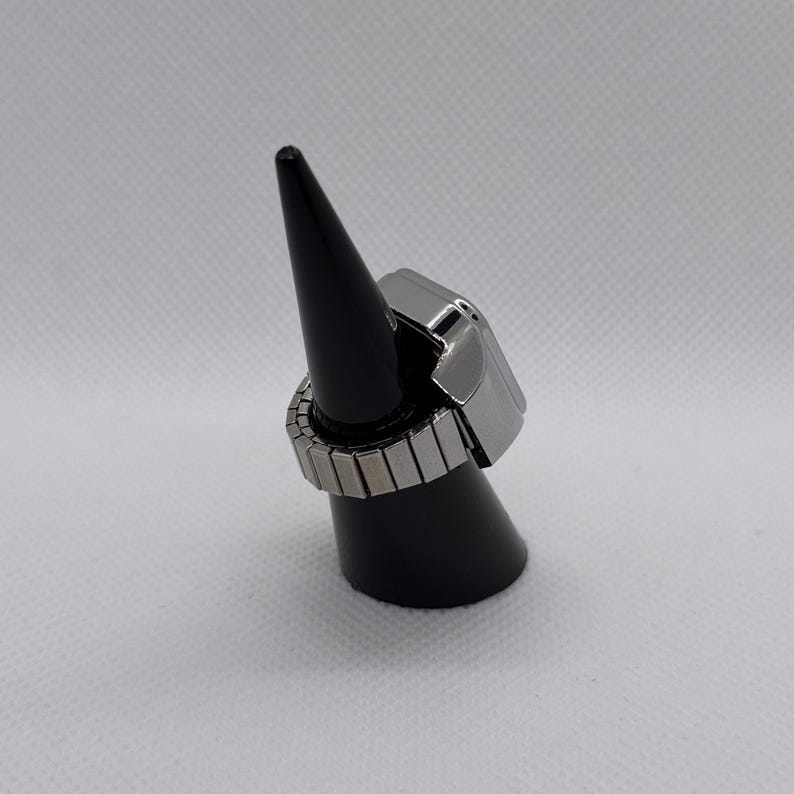 K&ouml;nnte beinhalten: Ein silberfarbener Ring mit rechteckiger Oberseite und verstellbarem Band. Der Ring wird auf einem schwarzen, kegelf&ouml;rmigen St&auml;nder vor wei&szlig;em Hintergrund pr&auml;sentiert. Das Band besteht aus kleinen, rechteckigen Metallst&uuml;cken.