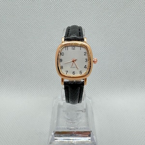 May include: A gold-toned quartz watch with a square face and black leather band. The watch face is white with black numerals and hands. The watch has a small crown on the right side.