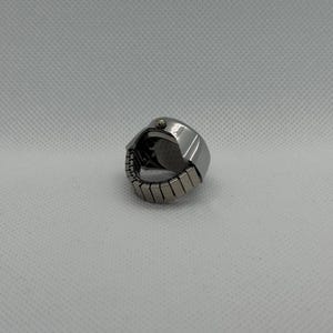 May include: A silver-toned ring watch with a round face and an adjustable band. The watch has a small crown and a dark watch face. The band is made of small, rectangular metal links.