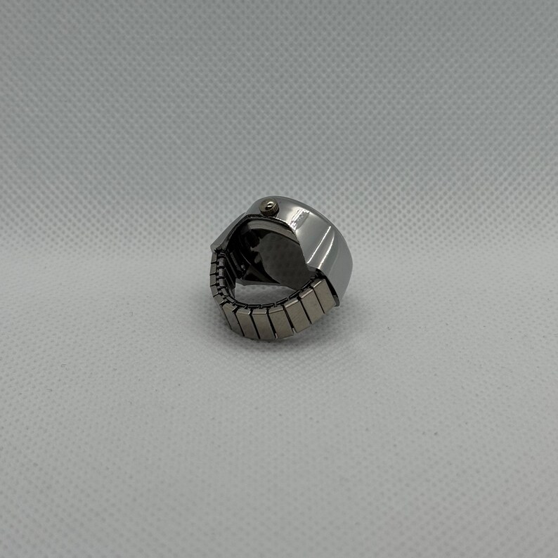 May include: A silver-toned ring watch with a round face and an adjustable band. The watch has a small crown and a dark watch face. The band is made of small, rectangular metal links.