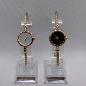 Puede incluir: Dos relojes de pulsera dorados con esferas redondas. Un reloj tiene una esfera blanca, el otro una esfera negra. Ambos tienen agujas doradas y un bisel dorado. Las correas tienen un diseño de cuerda trenzada. Los relojes se exhiben en soportes de plástico transparente.