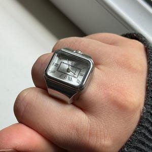 K&ouml;nnte beinhalten: Silberfarbener Uhrenring mit quadratischem Zifferblatt und r&ouml;mischen Ziffern. Das Zifferblatt ist hell und der Ring wird am Finger getragen. Der Ring ist ein einzigartiges Schmuckst&uuml;ck.