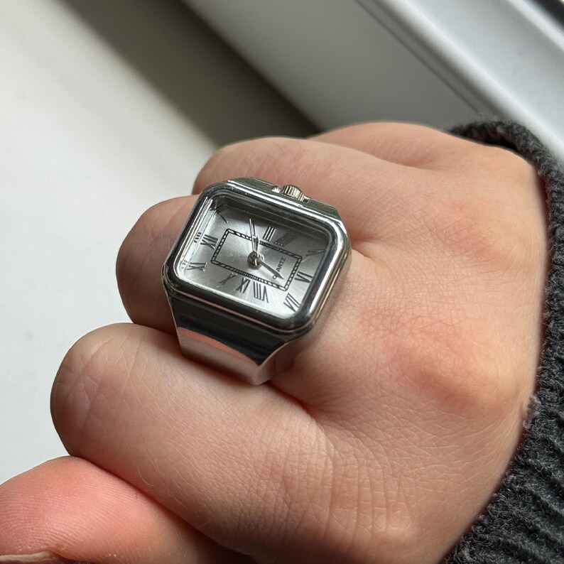 K&ouml;nnte beinhalten: Silberfarbener Uhrenring mit quadratischem Zifferblatt und r&ouml;mischen Ziffern. Das Zifferblatt ist hell und der Ring wird am Finger getragen. Der Ring ist ein einzigartiges Schmuckst&uuml;ck.