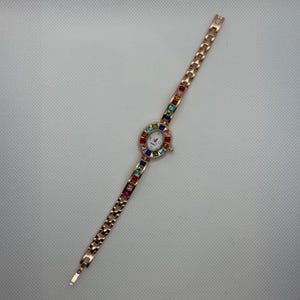 May include: A rose gold-toned wristwatch with a white face and colorful gemstone accents. The watch features a delicate chain-link band and a circular face surrounded by a rainbow of gemstones. The watch is designed for a small wrist.