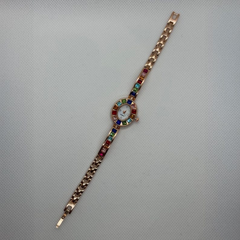 May include: A rose gold-toned wristwatch with a white face and colorful gemstone accents. The watch features a delicate chain-link band and a circular face surrounded by a rainbow of gemstones. The watch is designed for a small wrist.