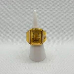 May include: A gold-toned quartz watch ring with a rectangular face. The watch face displays Roman numerals and hands. The ring is mounted on a white display stand.