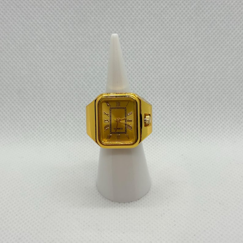 May include: A gold-toned quartz watch ring with a rectangular face. The watch face displays Roman numerals and hands. The ring is mounted on a white display stand.