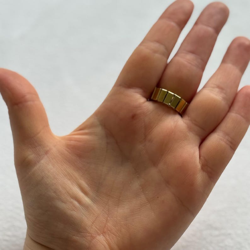 May include: A gold-colored ring with a rectangular design is worn on a finger. The ring's surface has a segmented appearance, with each segment appearing as a small, rectangular block. The ring is set against a neutral background.