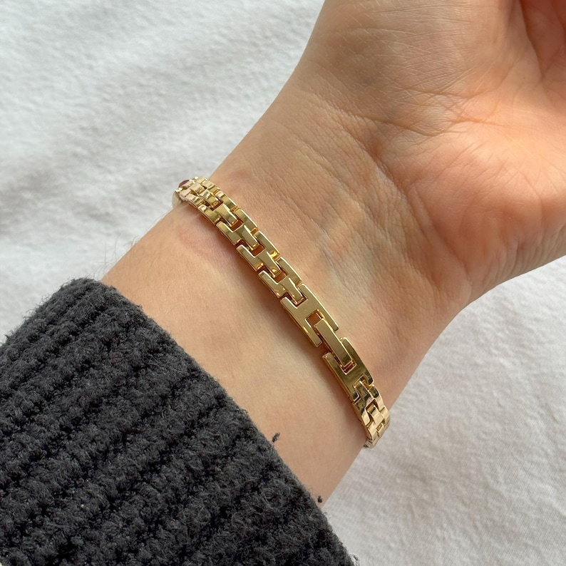 May include: A gold-colored bracelet with a rectangular link design. The bracelet is worn on a wrist, and the clasp is visible. The bracelet is made of a shiny, metallic material.