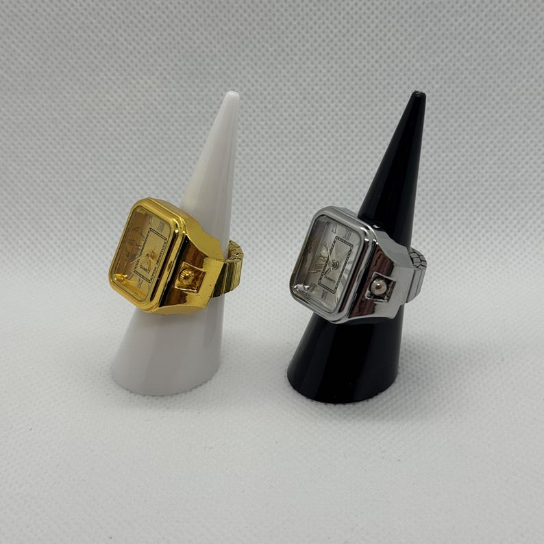 May include: Two ring watches, one gold-toned and one silver-toned, each with a rectangular watch face. The gold ring watch is on a white stand, and the silver ring watch is on a black stand. Both have an elastic band.