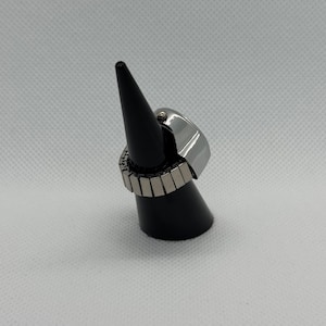 May include: A silver-toned ring with a rectangular face and an expandable band. The ring is displayed on a black cone-shaped stand against a white background. The band is made of small, rectangular metal pieces.