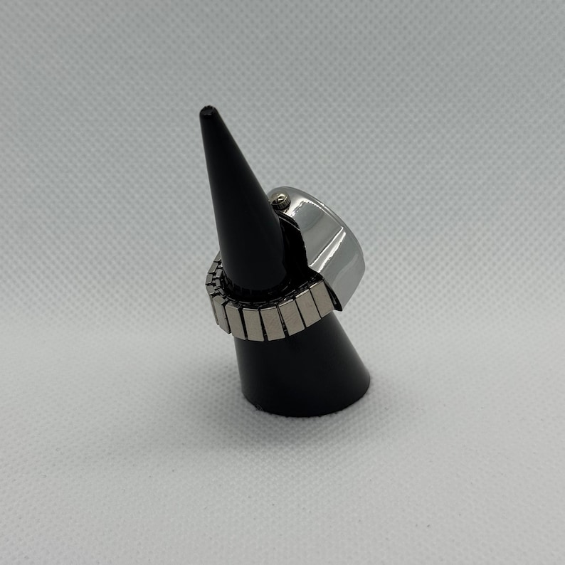 May include: A silver-toned ring with a rectangular face and an expandable band. The ring is displayed on a black cone-shaped stand against a white background. The band is made of small, rectangular metal pieces.