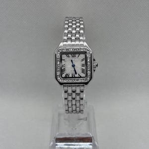 May include: A silver-tone quartz wristwatch featuring a square face with a textured white dial, black Roman numeral hour markers, and blue hands. The bezel is encrusted with small clear stones, and the watch has a multi-link metal bracelet.