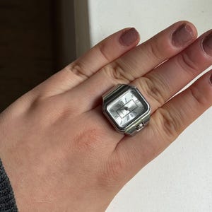 K&ouml;nnte beinhalten: Eine silberfarbene Ringuhr mit rechteckigem Zifferblatt und r&ouml;mischen Ziffern. Die Uhr hat ein silbernes Band und eine kleine Krone an der Seite. Die Uhr wird am Finger getragen.