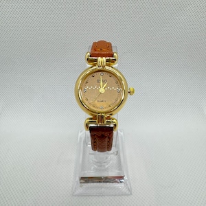 Peut inclure: Une montre à quartz dorée avec un bracelet en cuir marron. Le cadran de la montre est de couleur beige clair avec de petits diamants et le texte "XS VO QUARTZ". La montre est présentée sur un présentoir en plastique transparent.