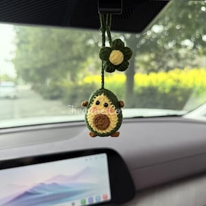May include: A handmade crochet car charm featuring an avocado design with a smiling face and a flower. The avocado is green, brown, and yellow, with small arms and legs. The flower is green with a yellow center. The charm hangs from a dark green string.