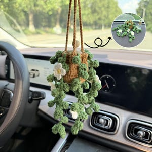 Könnte beinhalten: Ein handgefertigter gehäkelter Autoanhänger mit einem hängenden Pflanzendesign. Der Anhänger hat einen braunen Topf, grüne Blätter und weiße Blüten. Er wird von einer braunen Kordel und einer Holzperle gehalten, um dem Auto-Innenraum einen Hauch von Natur zu verleihen.