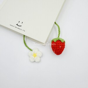 May include: A handmade crochet bookmark featuring a red strawberry and a white flower with a yellow center. The bookmark is attached to a book with the text "record the beauty life." The bookmark is a charming accessory for book lovers.