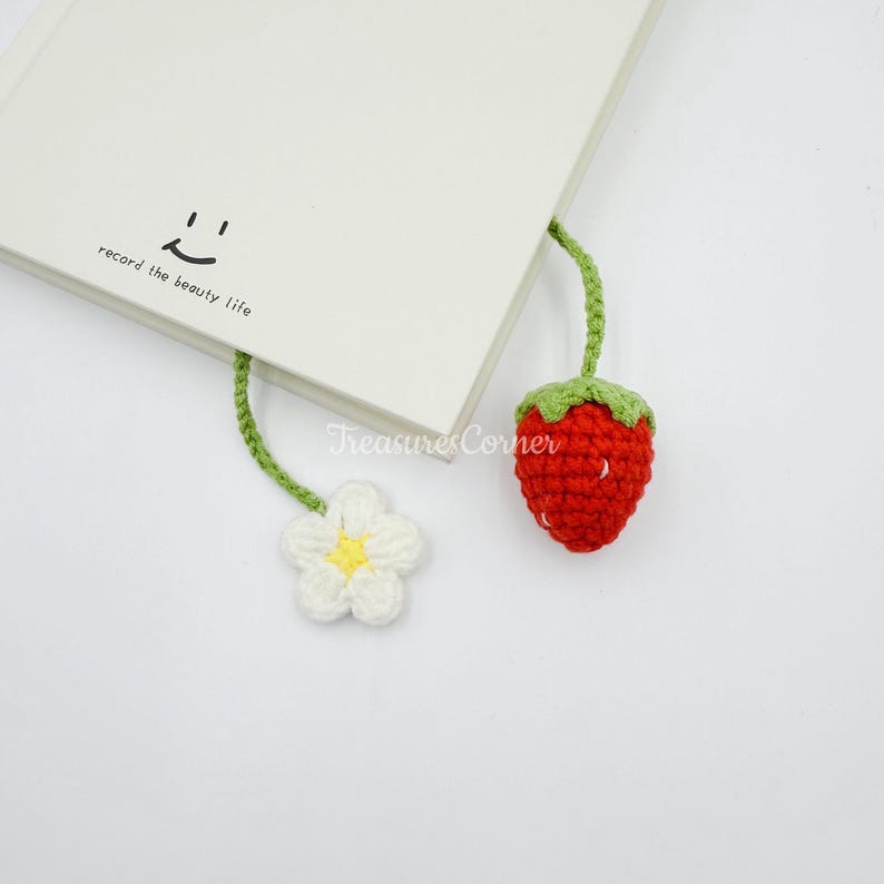 May include: A handmade crochet bookmark featuring a red strawberry and a white flower with a yellow center. The bookmark is attached to a book with the text "record the beauty life." The bookmark is a charming accessory for book lovers.