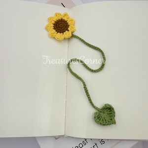 May include: A handmade crochet sunflower bookmark with a yellow flower, brown center, and green stem and leaf. The bookmark is placed on an open book. The text "TreasuresCorner" is visible.