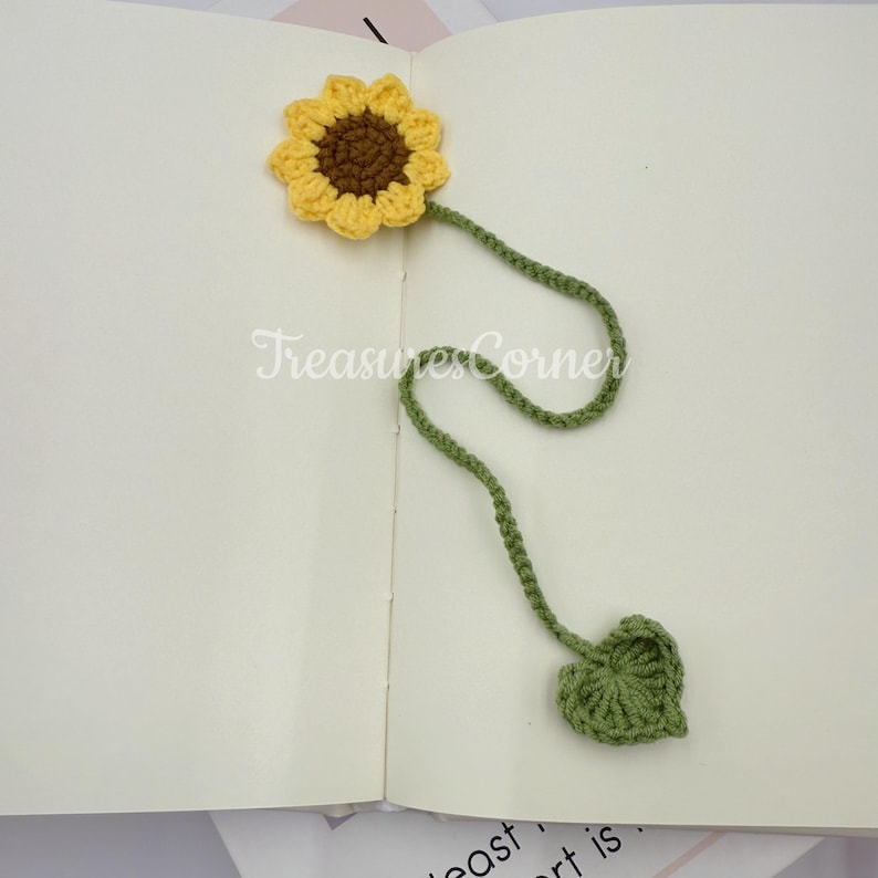 May include: A handmade crochet sunflower bookmark with a yellow flower, brown center, and green stem and leaf. The bookmark is placed on an open book. The text "TreasuresCorner" is visible.