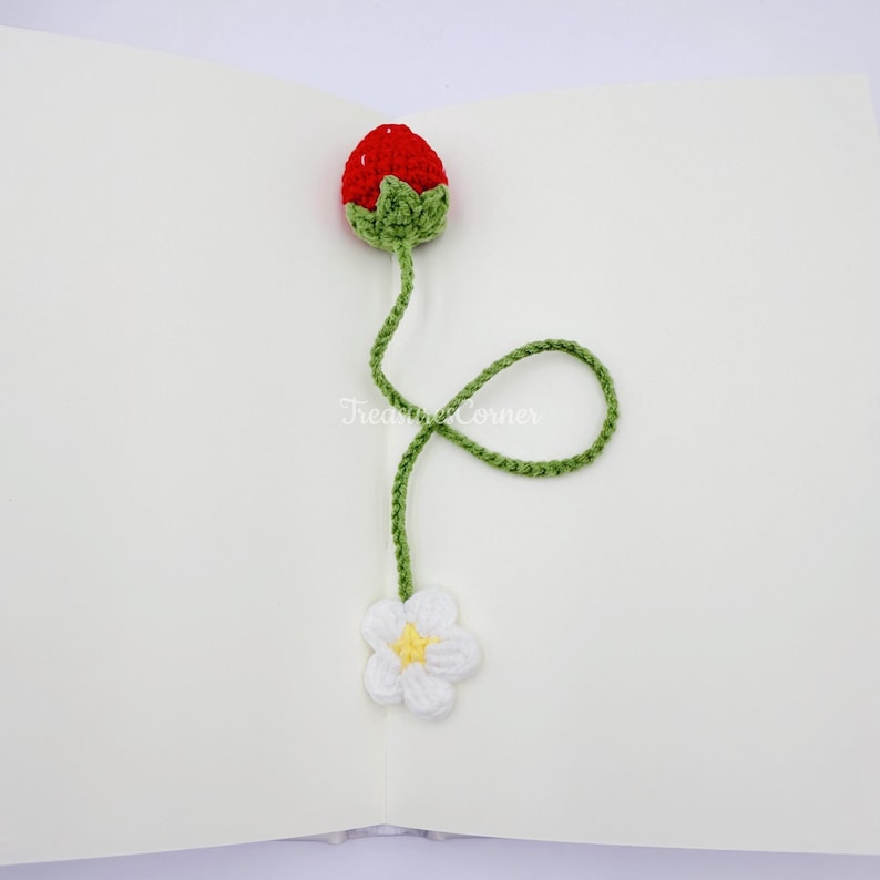 May include: Crocheted strawberry bookmark with a red berry, green leaves, and a green stem that curls down to a white flower with a yellow center. The bookmark is handmade and rests on a white surface.
