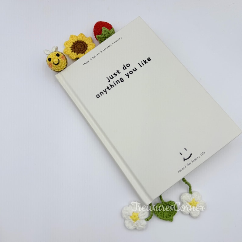May include: A white book with the text "just do anything you like" and a smiling face. Crochet bookmarks in the shape of a bee, sunflower, strawberry, and flowers are placed in the book. The text "TreasuresCorner" is visible.