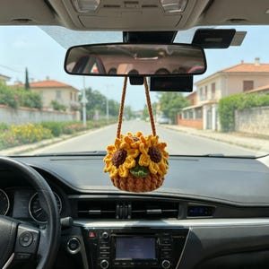 May include: A handmade crochet sunflower car charm. The charm features a yellow sunflower design with brown and green accents, suspended from a brown cord. The charm is hanging from the rearview mirror inside a car.