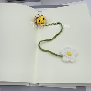 May include: A handmade crochet bookmark featuring a yellow and brown bee with white wings, a green stem, and a white flower with a yellow center. The text "TreasuresCorner" is visible on the page.