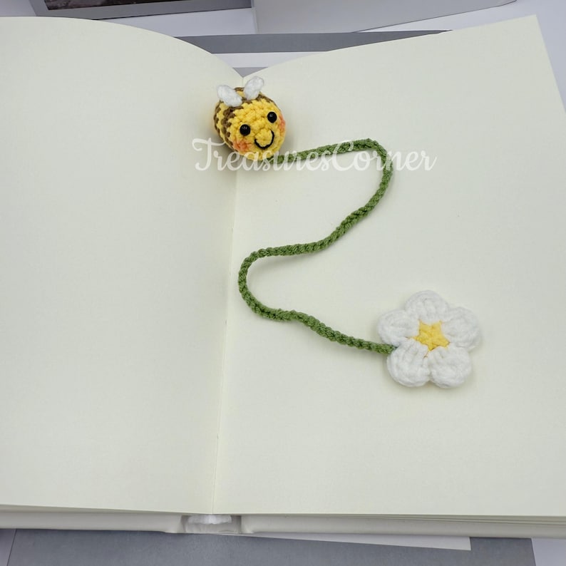 May include: A handmade crochet bookmark featuring a yellow and brown bee with white wings, a green stem, and a white flower with a yellow center. The text "TreasuresCorner" is visible on the page.