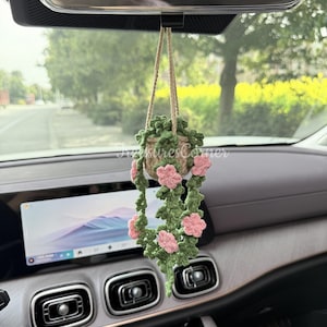 May include: A crocheted hanging plant featuring pink flowers and green leaves, suspended by a beige string. The plant is in a woven basket. The item is hanging from a car's rearview mirror.