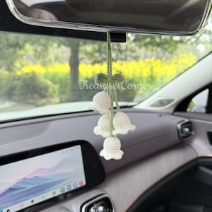 Puede incluir: Un adorno de coche de crochet blanco con forma de lirio de los valles, colgado del espejo retrovisor de un coche. El adorno presenta cuatro flores en forma de campana y un lazo verde. El texto "TreasuresCorner" es visible.