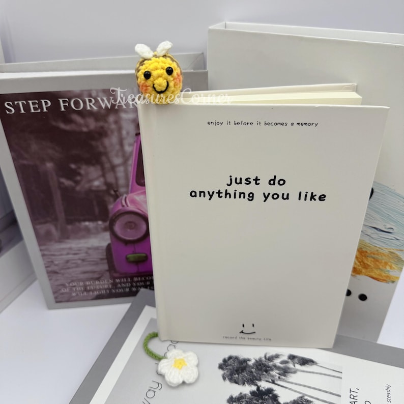 May include: A crocheted bee bookmark rests on a white notebook with the text "just do anything you like." A white flower bookmark and a book with the text "STEP FORWARD" are also visible.