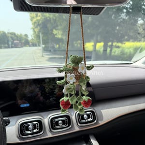 Pode incluir: Um pendente de carro artesanal com um sapo de crochê em uma cesta suspensa com flores brancas e morangos vermelhos. O pendente é suspenso por uma corda marrom.