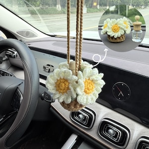 May include: A crocheted daisy flower car air freshener hanging from the rearview mirror. The flowers have white petals and yellow centers, set in a brown basket. A small glass bottle with a wooden top is also visible.