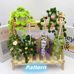 May include: Five crocheted hanging plant pots with green, pink, and purple leaves. The pots are hanging from a wooden shelf with the word "Pattern" written on the bottom.