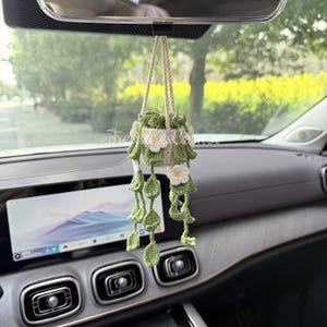May include: A handmade crochet car charm featuring a small hanging planter with green leaves and white flowers. The planter is suspended by a beige cord and hangs from a car's rearview mirror. The charm adds a touch of nature to the car's interior.