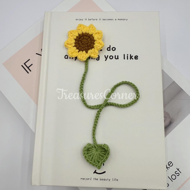 May include: A handmade crochet sunflower bookmark with a yellow flower, brown center, and green stem and leaf. The bookmark is placed on a white book with text that reads "enjoy it before it becomes a memory".
