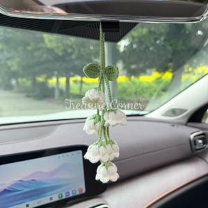 Pode incluir: Um pendente de carro feito à mão, de crochê, com flores brancas de lírio-do-vale com caules e folhas verdes. O pendente está suspenso do espelho retrovisor de um carro. O texto "Treasure Corner" é visível.