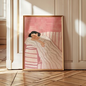 May include: A framed art print depicts a person sleeping on a striped sofa, draped in a blanket. The artwork features a soft color palette of pink, white, and gray, with a gold-colored frame. The illustration evokes a sense of calm and relaxation.